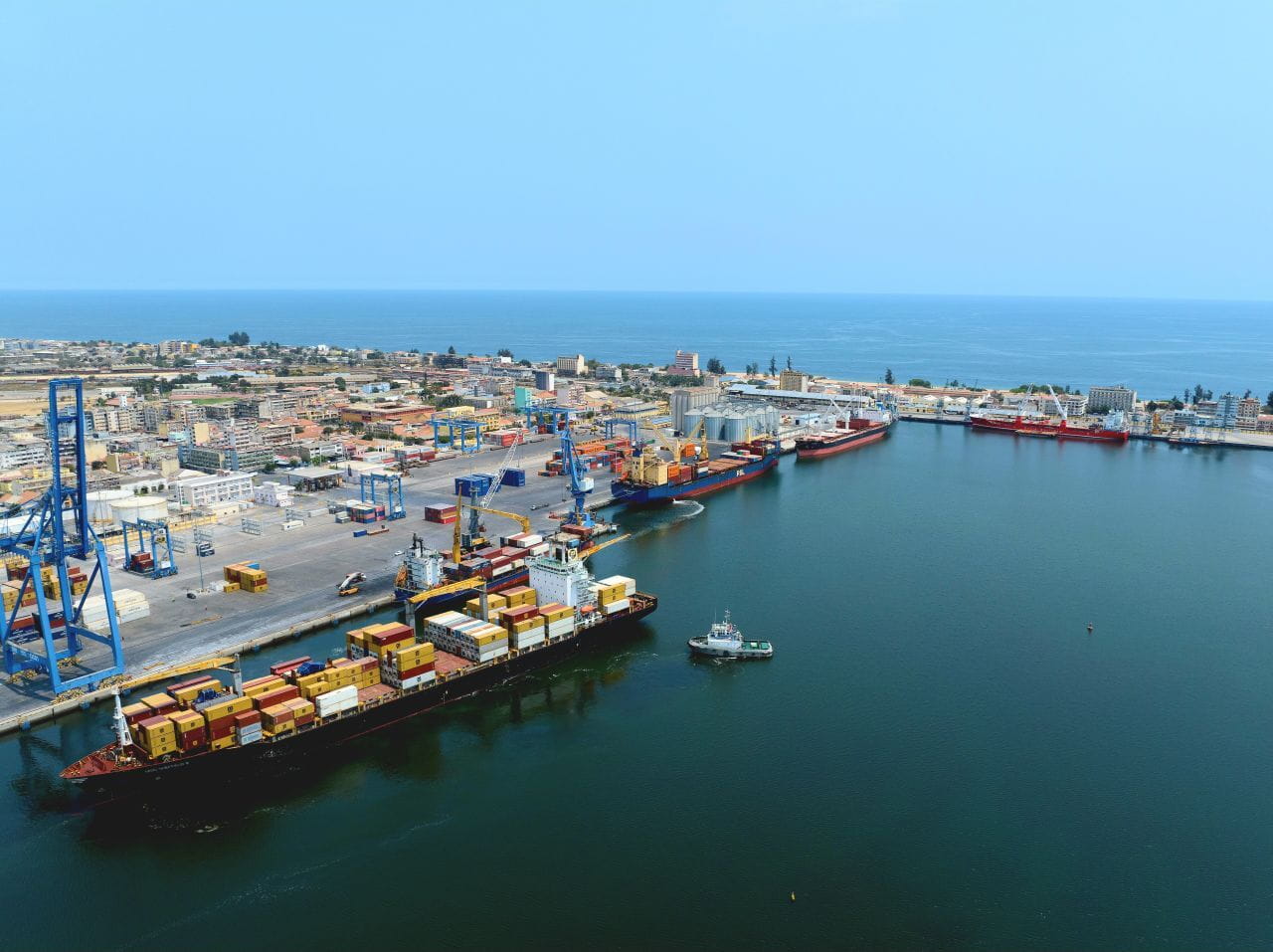 In a view of the AGL Lobito Terminal, a large ship floats beside a port, loaded with multiple shipping containers of various colors. 