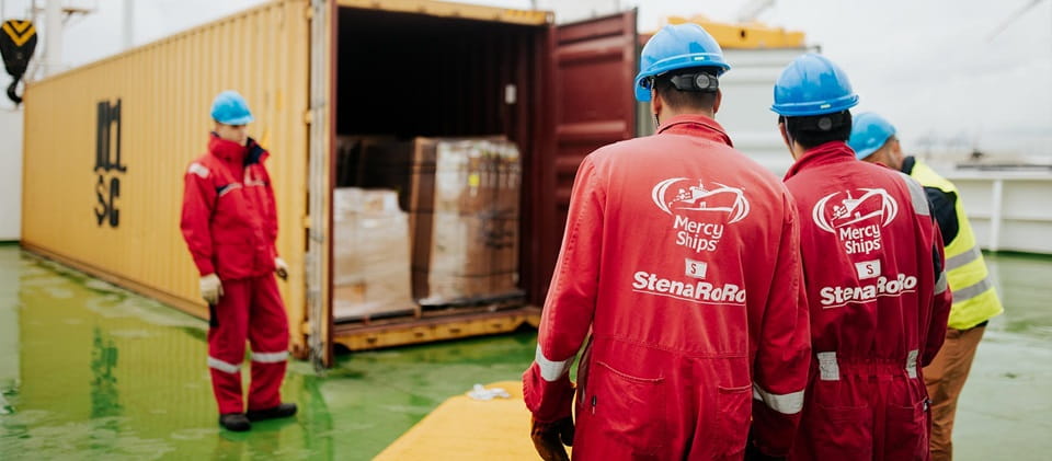 Three men in red work clothes stand beside a large container, engaged in conversation and teamwork.